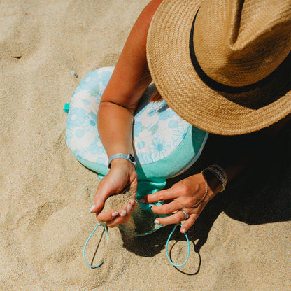 Overhead view of a woman in a straw hat sitting on sand, wearing bracelets and rings, leaning against The Darling Effect Daisy Craze Aqua Ballast Beach Pillow as she lets sand fall through her hands.