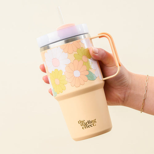 A hand holds the Daisy Craze Peach 20 oz Mini On-The-Go Tumbler by The Darling Effect, featuring a beige base with pastel daisies, a handle, and a clear lid with straw—perfect for stylish hydration on the go.