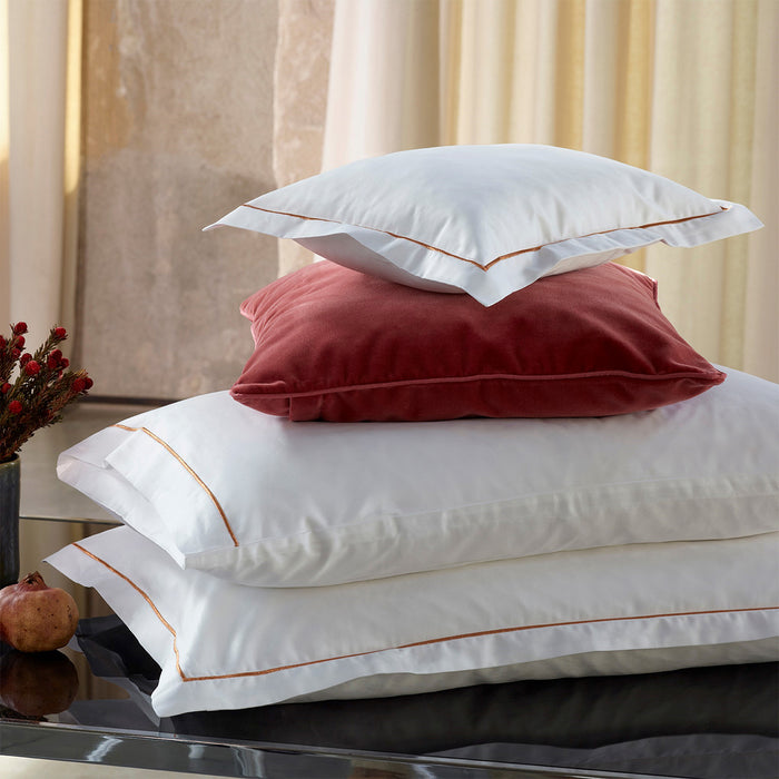 Stack of Dalia white/rose pillowcase sham on a bed with beige curtains.
