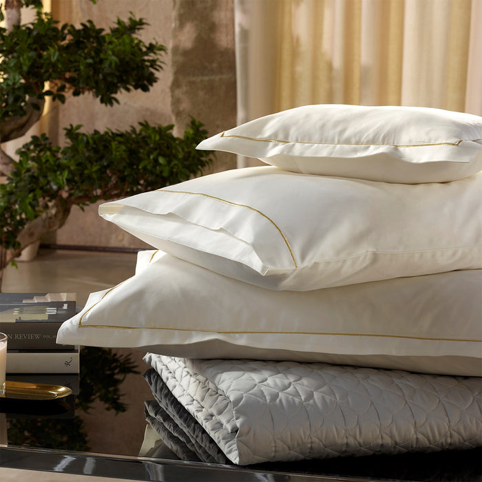 Stack of Dalia cream/gold pillowcase sham on a reflective surface, with a blurred indoor setting in the background.