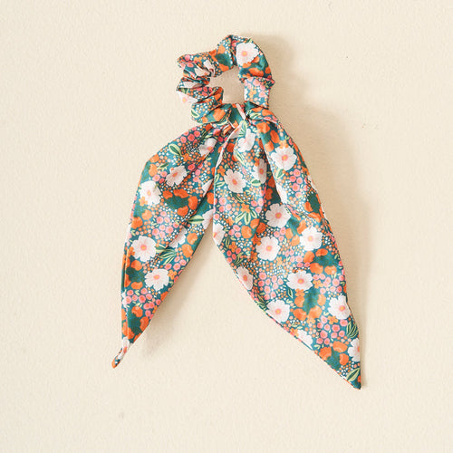 The Darling Effect’s Dark Green with scrunchie Sweet Meadow Printed Hair Scarf features pink, white, and orange floral prints on a dark green background, displayed on a light beige surface.