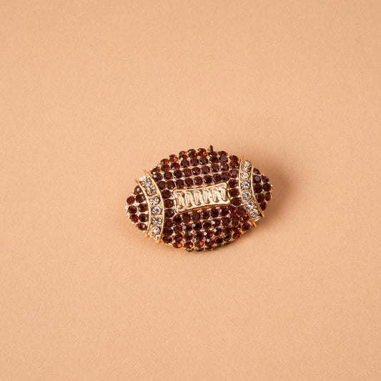 The Dazzle Football Timeless Brooch by The Darling Effect, a vintage-inspired accessory shaped like an American football and adorned with brown and clear rhinestones, rests on a plain beige background. This keepsake statement piece exudes classic charm and unique style.