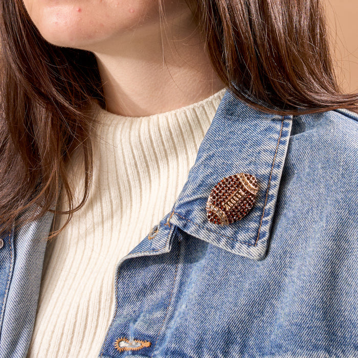A person wearing a cream ribbed sweater and a blue denim jacket with The Darling Effect’s Dazzle Football Timeless Brooch—a vintage-inspired, beaded pin—in the shape of a football attached to their lapel. Only the lower half of their face and hair are visible.