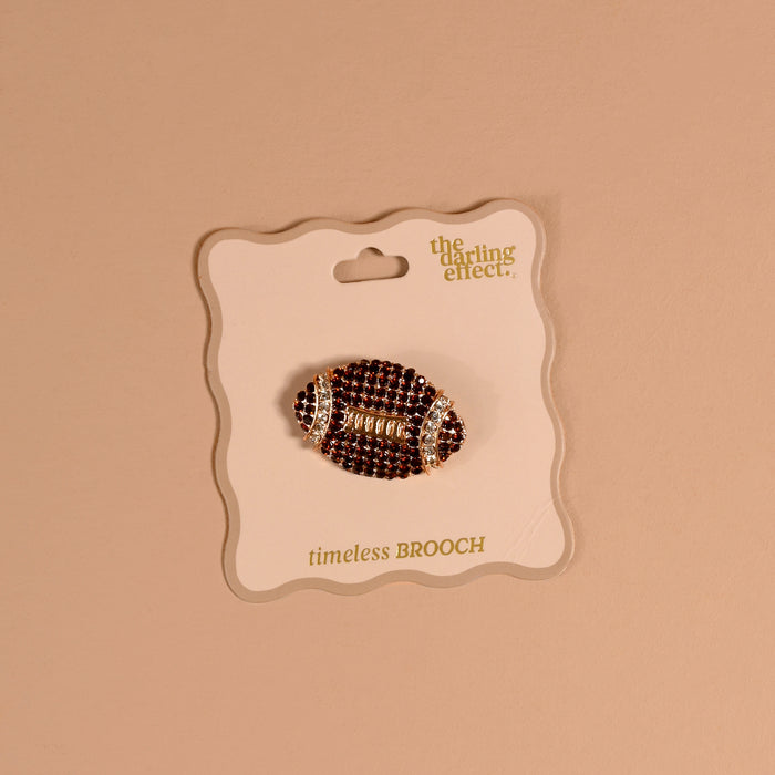 The Dazzle Football Timeless Brooch by The Darling Effect, featuring a vintage-inspired rhinestone football design, is attached to a card labeled “the darling effect. timeless BROOCH” and displayed against a beige background—making it a charming keepsake accessory.