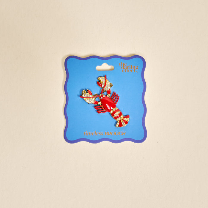 The Darling Effect’s Dazzle Lobster Timeless Brooch, in red and gold and presented on a blue “timeless BROOCH” card, shines as an elegant keepsake accessory against a light beige background.