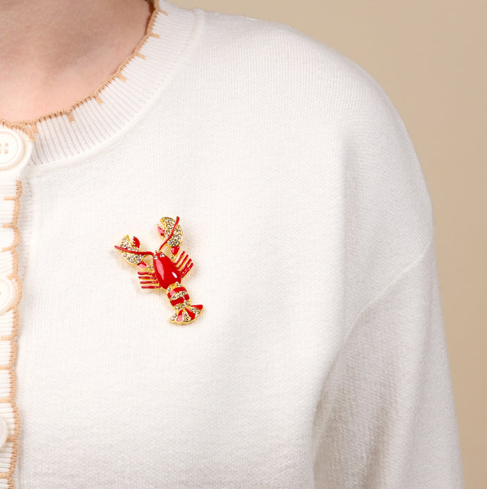 A person wears a white button-up sweater with The Darling Effect's Dazzle Lobster Timeless Brooch—an elegant vintage-inspired red and gold lobster pin—on the left shoulder.