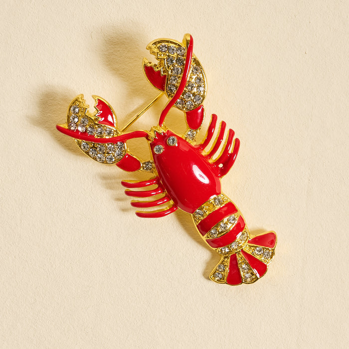 The Darling Effect’s Dazzle Lobster Timeless Brooch features vintage-inspired gold with red enamel and rhinestone accents on the claws and tail, displayed on light beige—a charming keepsake accessory.