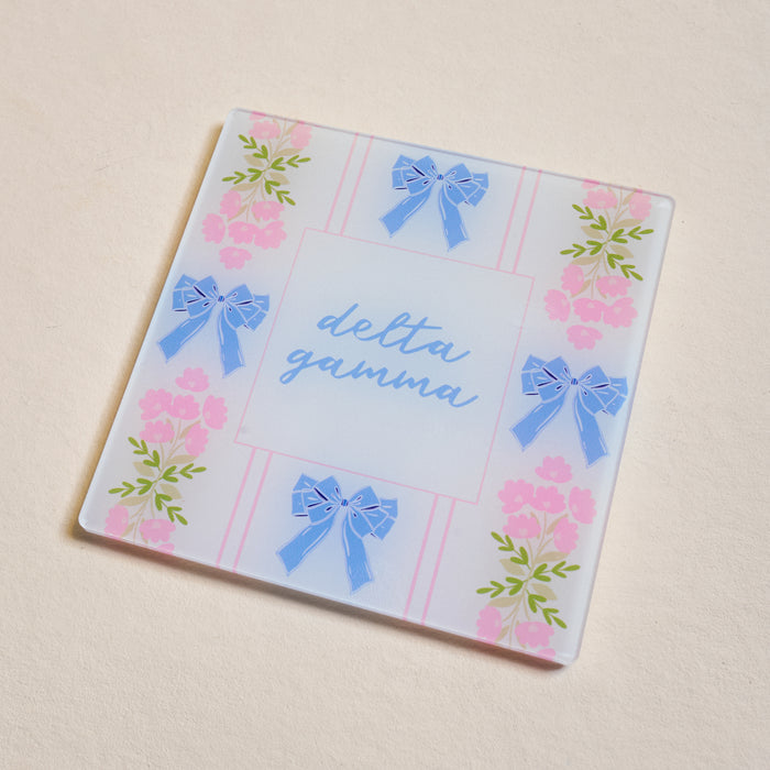 The Darling Effect’s Delta Gamma Acrylic Coaster features blue script, blue bows, and pink flowers on a pastel background—an ideal and charming Big/Little gift for any sorority member.