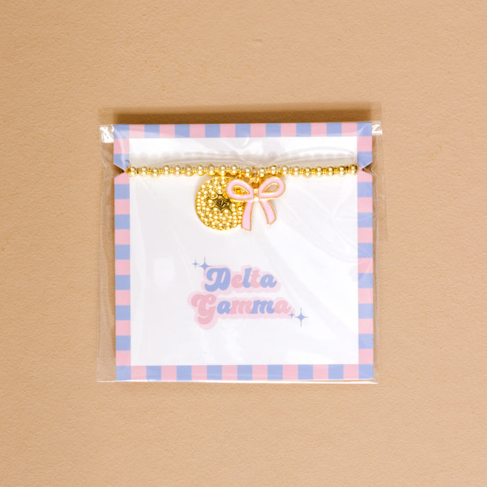 The Darling Effect's Delta Gamma Bow Gold Charm Bracelet features a gold beaded design with a circular Delta Gamma charm and pink bow, packaged on a pastel plaid card—an ideal sorority gift or stylish jewelry piece.