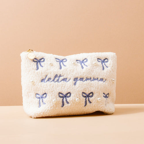 The Delta Gamma Bows & Pearls Teddy Pouch by The Darling Effect features fluffy white sherpa, blue embroidered bows and pearls, and "delta gamma" in blue script—making it a perfect sorority gift.