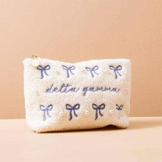 The Delta Gamma Bows & Pearls Teddy Pouch by The Darling Effect features fluffy white sherpa, blue embroidered bows and pearls, and "delta gamma" in blue script—making it a perfect sorority gift.