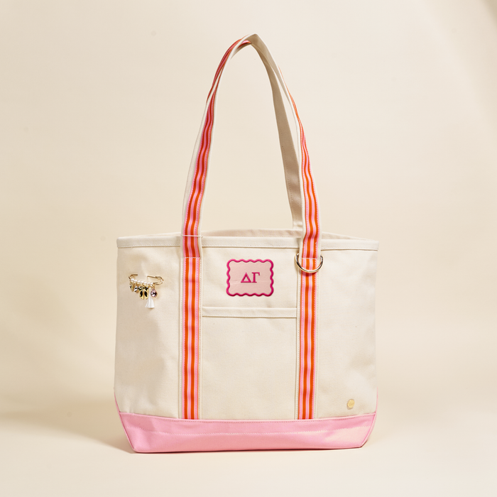 The Darling Effect’s Delta Gamma Canvas Tote Bag features beige canvas, pink accents, striped handles, a front patch with Greek letters ΔΓ trimmed in pink, and a gold keychain—an ideal sorority gift. Light beige background.