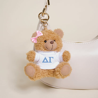 The Darling Effect’s Delta Gamma Teddy Bear Bag Charm features a small brown bear with a pink bow and white shirt with Greek letters, hanging from a gold keychain—ideal for accessorizing your white handbag.