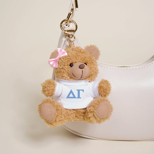 The Darling Effect’s Delta Gamma Teddy Bear Bag Charm features a small brown bear with a pink bow and white shirt with Greek letters, hanging from a gold keychain—ideal for accessorizing your white handbag.