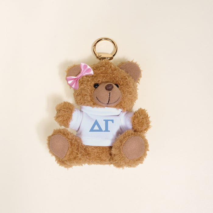 The Darling Effect's Delta Gamma Teddy Bear Bag Charm is a small brown teddy keychain with a pink bow, wearing a white shirt featuring blue ΔΓ Greek letters—perfect as a sorority gift or cute accessory.