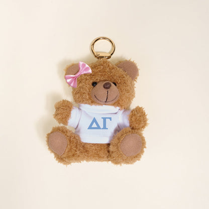The Darling Effect's Delta Gamma Teddy Bear Bag Charm is a small brown teddy keychain with a pink bow, wearing a white shirt featuring blue ΔΓ Greek letters—perfect as a sorority gift or cute accessory.