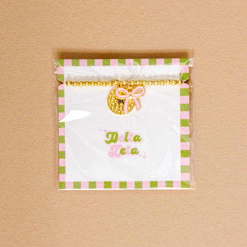 Show your chapter pride with The Darling Effect’s Delta Zeta Bow Gold Charm Bracelet, featuring gold beads, a pink bow, and displayed on a green and pink checkered card.
