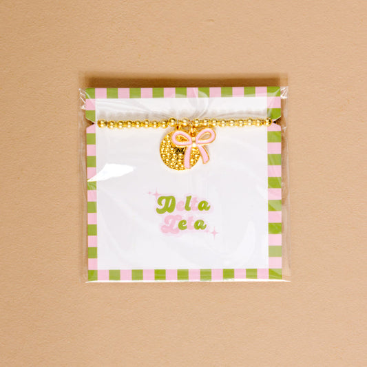 Show your chapter pride with The Darling Effect’s Delta Zeta Bow Gold Charm Bracelet, featuring gold beads, a pink bow, and displayed on a green and pink checkered card.