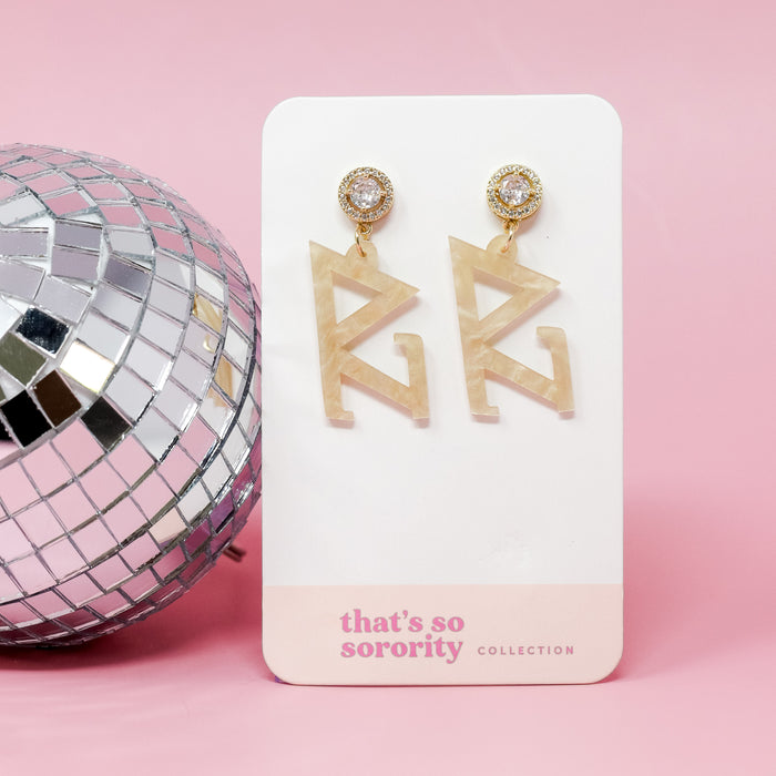 A pair of Delta Zeta Letter Gold Acrylic Earrings by The Darling Effect is displayed on a white card, with a disco ball beside it—perfect for bid-day baskets or sisterhood events—all set against a pink background.