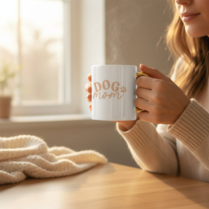 A woman in a beige sweater enjoys coffee from The Darling Effect Dog Mom 11 oz Cozy Cute Ceramic Mug with gold lettering and handle, sitting at a sunlit wooden table wrapped in a blanket as steam rises from her drink.
