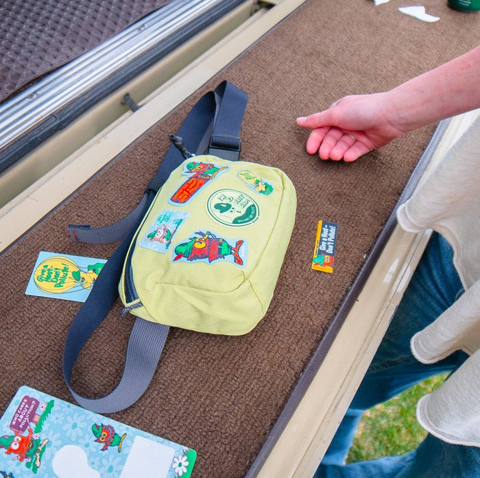 Yellow fanny pack with NoSo Don’t Pollute Woodsy Owl patch beside playing cards and an outstretched arm, eco-friendly style.