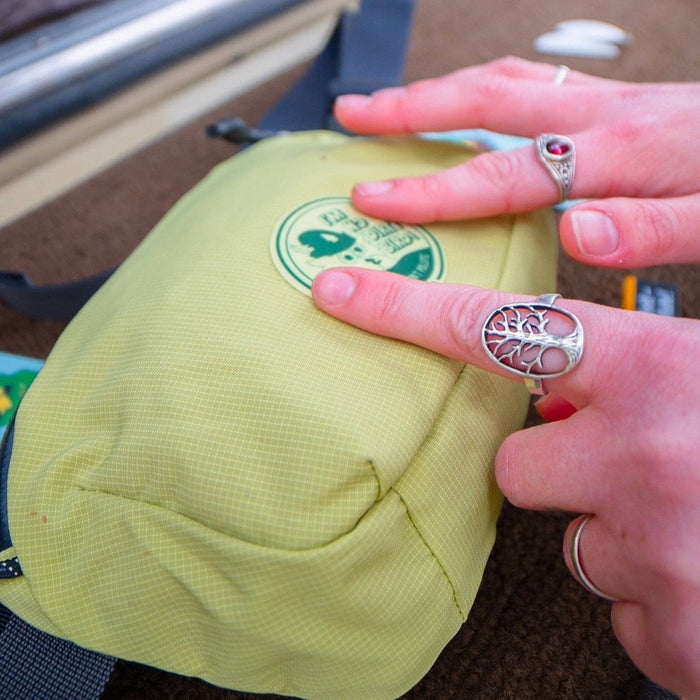 Hands with silver rings point to a Woodsy Owl NoSo Repair Patch on a yellow Noso Patches bag; eco-friendly gear repair.