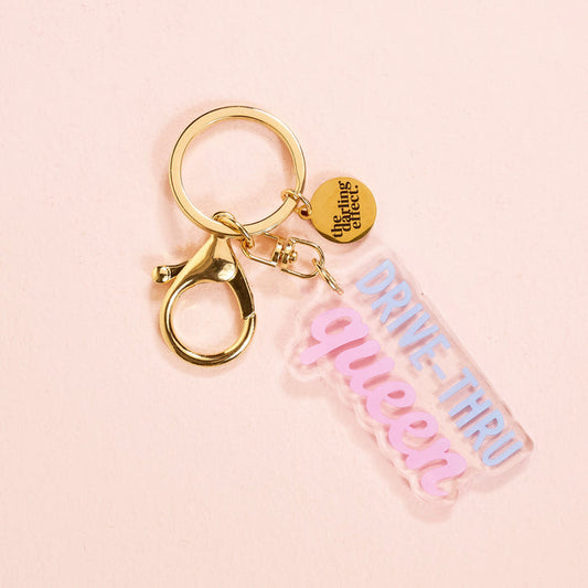 The Drive Thru Queen Acrylic Keychain by the-darling-effect features a gold keyring, a small round tag, and a clear acrylic tag with "DRIVE-THRU queen" in blue and pink—perfect for cute keychain lovers. Shown on a light pink background.