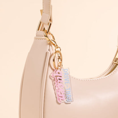 The Drive Thru Queen Acrylic Keychain by the-darling-effect, in pastel pink and blue, hangs from a beige shoulder bag—an adorable accessory for fans of cute keychains.