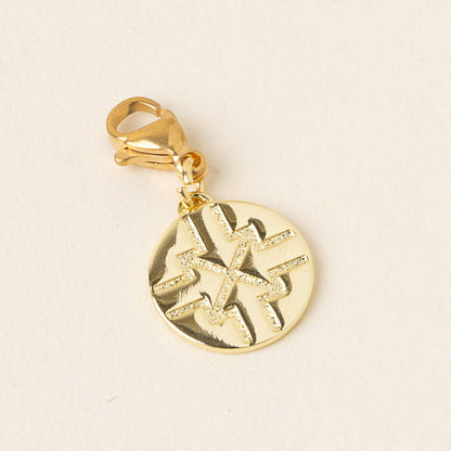 The Driven Jewelry Trait Charm Gold by the-darling-effect features a round gold charm with an abstract geometric engraving and a lobster clasp, shown on a light, neutral background.