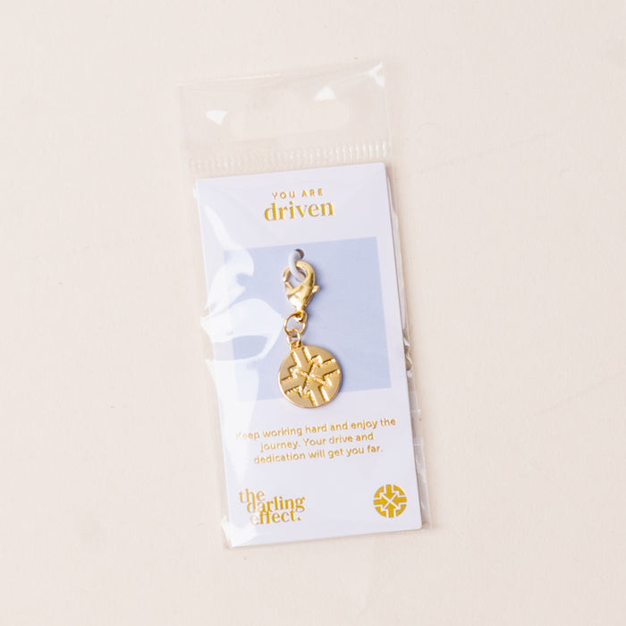 The Driven Jewelry Trait Charm Gold by the-darling-effect features a gold compass charm on a lobster clasp, presented on a card that encourages drive, hard work, and enjoying the journey.