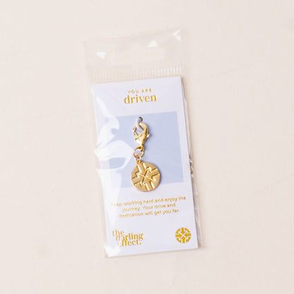The Driven Jewelry Trait Charm Gold by the-darling-effect features a gold compass charm on a lobster clasp, presented on a card that encourages drive, hard work, and enjoying the journey.