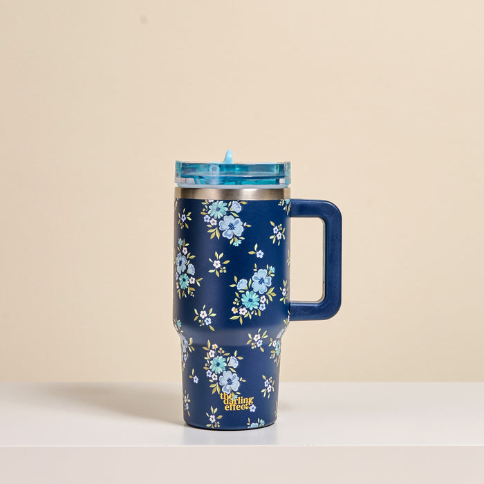 A navy Endless Daydream 20 oz Lifestyle Flip Straw Tumbler by the-darling-effect with a handle, blue lid, and light blue floral pattern rests on a white surface against a beige background. The Little Clinic logo appears near the base.