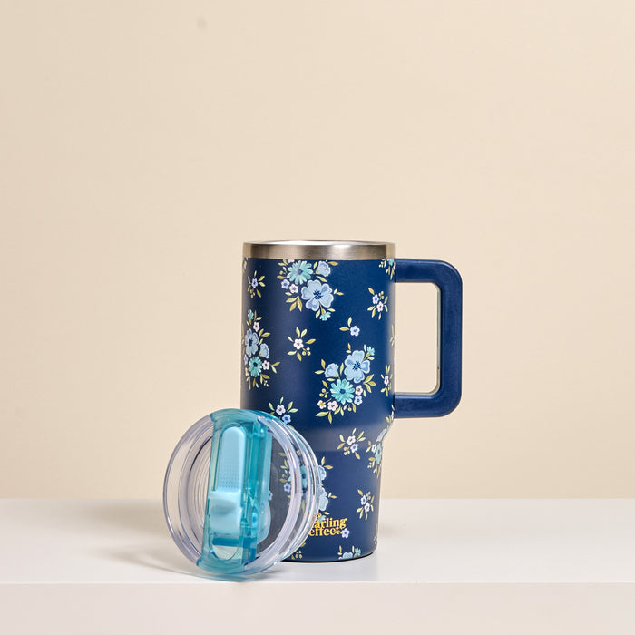 The-darling-effect Endless Daydream Navy 20 oz Lifestyle Flip Straw Tumbler stands upright with its clear lid featuring a blue sliding cover resting beside it. The insulated stainless steel tumbler has light blue floral patterns.