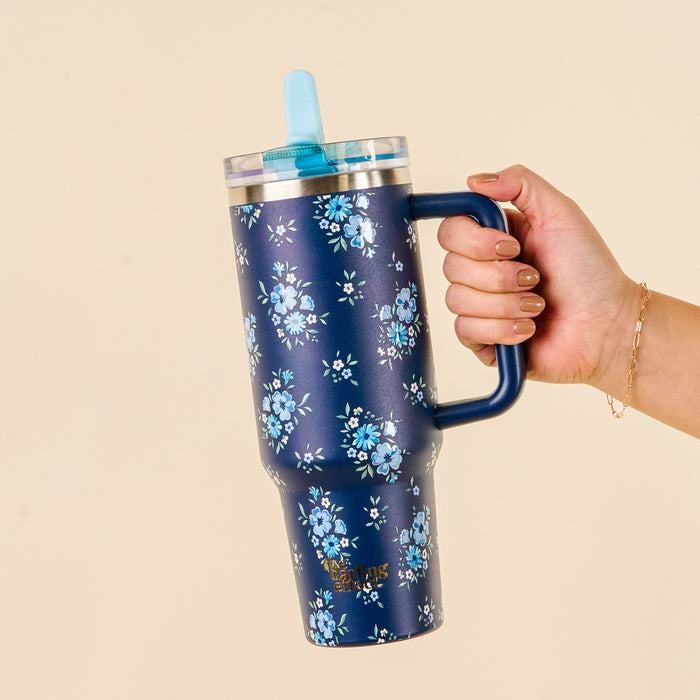 A hand holds the-darling-effect Endless Daydream Navy 40 oz Lifestyle Flip Straw Tumbler, featuring a blue floral pattern, clear lid, blue straw, and handle. Manicured nails and a gold bracelet add style against a beige background.