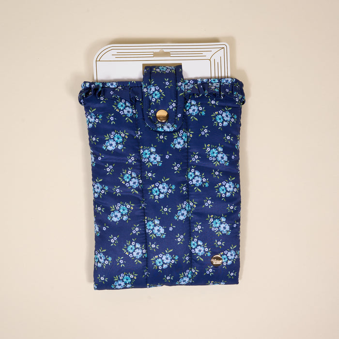The Darling Effect’s Endless Daydream Navy Book Cover features quilted navy fabric with a blue floral print, ruffled top edge, gold snap, and logo charm. Doubles as a book sleeve. Shown on a white hanger tag against a beige background.