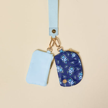 The-darling-effect’s Endless Daydream Navy Dynamic Duo Pouch Wristlet features a pale blue lanyard with gold clasp and two zippered pouches—a solid light blue and a floral navy—combining hands-free luxe style against a beige background.