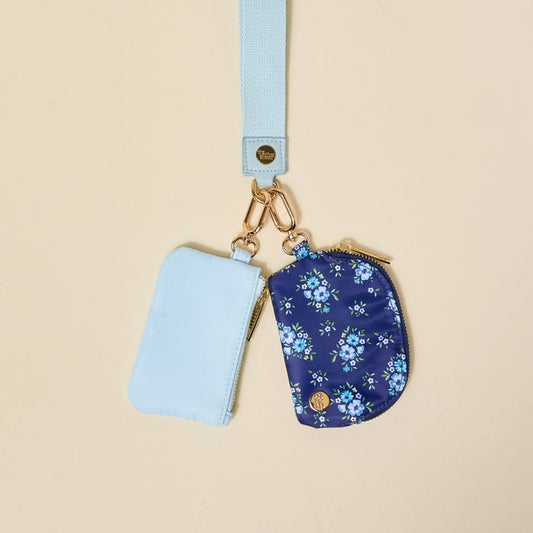 The-darling-effect’s Endless Daydream Navy Dynamic Duo Pouch Wristlet features a pale blue lanyard with gold clasp and two zippered pouches—a solid light blue and a floral navy—combining hands-free luxe style against a beige background.