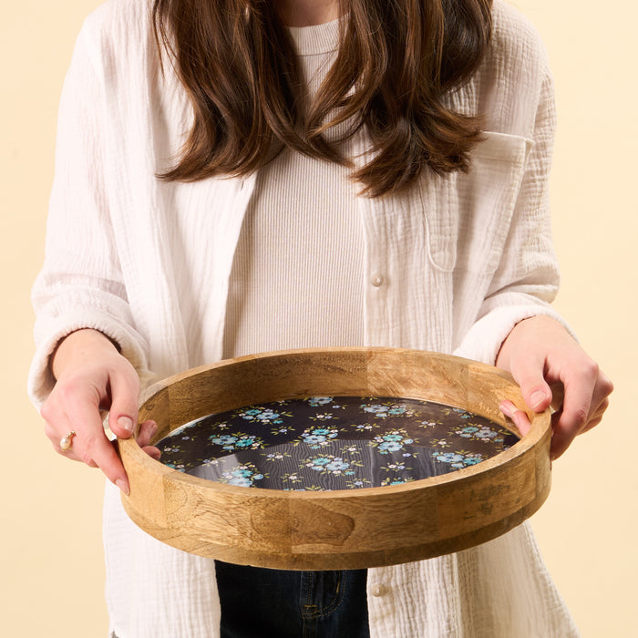 A woman in a white cardigan holds the-darling-effect's Endless Daydream Navy Large Round Wood Enamel Tray, featuring a dark floral interior with blue and white flowers on a black background.