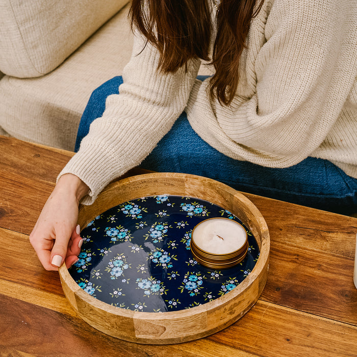 A person's hand rests on the-darling-effect Endless Daydream Navy Large Round Wood Enamel Tray, which features a blue floral pattern. A candle sits in the serving tray.
