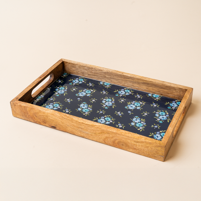 The-darling-effect’s Endless Daydream Navy Rectangle Wood Enamel Tray features cutout handles and a blue floral pattern on a dark base. Food-safe, it rests elegantly on any beige surface.