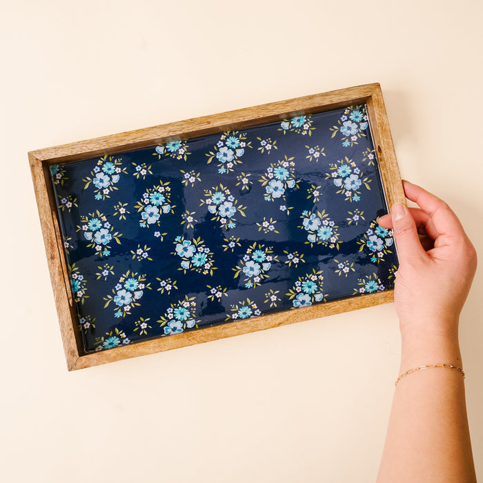 A hand holds the Endless Daydream Navy Rectangle Wood Enamel Tray by the-darling-effect, featuring a blue floral pattern on a dark background, against a light beige backdrop.