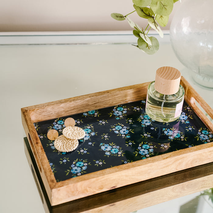 The-darling-effect’s Endless Daydream Navy Rectangle Wood Enamel Tray holds gold earrings and a wooden-capped perfume bottle. It rests on glass next to a vase of green stems.