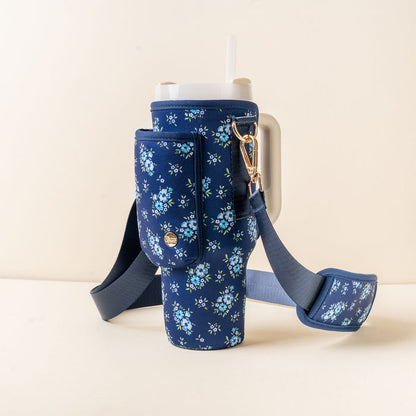 The-darling-effect’s Endless Daydream Navy Tumbler Sling, featuring a blue floral pattern, zippered pocket, and adjustable hands-free strap, holds a large tumbler with handle and straw against a plain light background.