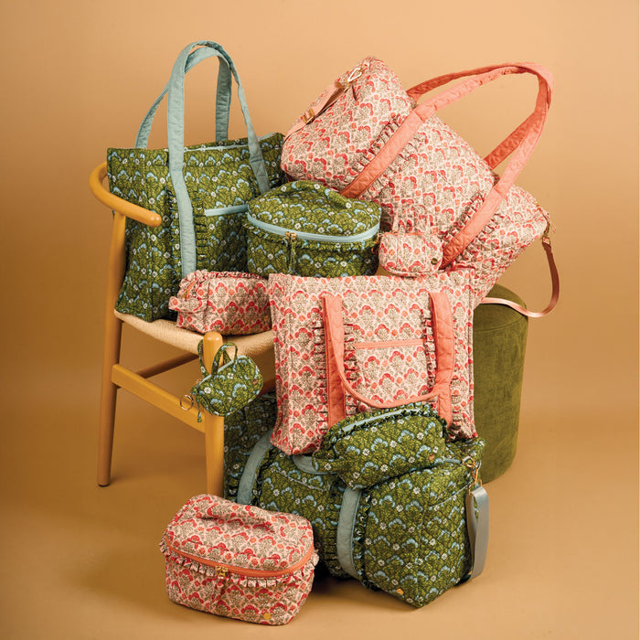 The Darling Effect’s Everbloom Dusty Rose Glam & Go Quilted Cosmetic Bag is displayed with totes, backpacks, pouches, and round cases in green and pink floral patterns on and around a wooden chair against a beige background.