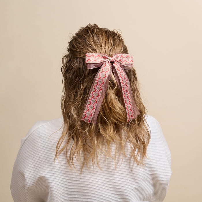 Seen from behind against a beige background, a person with wavy light brown hair wears a white top and The Darling Effect's Everbloom Dusty Rose Lovely Layered Hair Bow, featuring a pink patterned design tied in their hair.