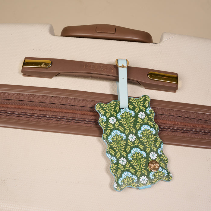 The Darling Effect Everbloom Forest Green Jetsetter Luggage Tag, featuring a green and blue floral pattern, privacy flap, and gold label, is attached to a beige Delsey suitcase with brown trim—an ideal travel accessory.