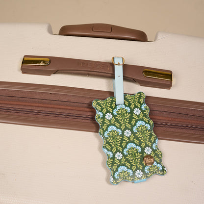The Darling Effect Everbloom Forest Green Jetsetter Luggage Tag, featuring a green and blue floral pattern, privacy flap, and gold label, is attached to a beige Delsey suitcase with brown trim—an ideal travel accessory.