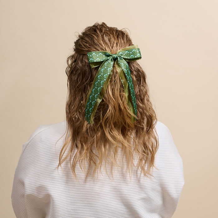 A woman’s wavy blonde hair is styled with The Darling Effect’s Everbloom Forest Green Lovely Layered Hair Bow—an elegant, floral-inspired accessory choice.