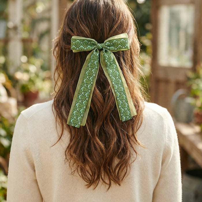 Back view of a woman with brown wavy hair styled half-up, secured with The Darling Effect's Everbloom Forest Green Lovely Layered Hair Bow. She wears a cream sweater and displays this charming accessory in a garden setting.