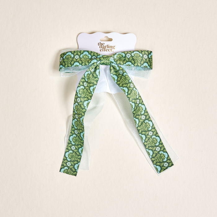 The Everbloom Forest Green Lovely Layered Hair Bow by The Darling Effect features long tails and comes on white packaging, ideal for stylish hair accessory lovers. Set against a light beige background.